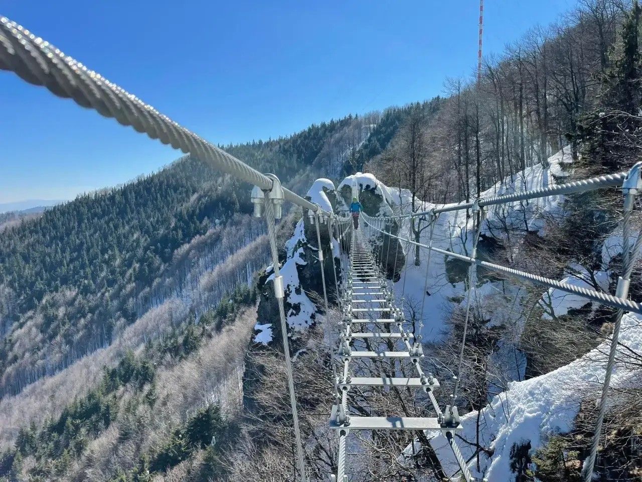 Osoba na moście wiszącym pokonuje trasę ferrata, podziwiając zimowy krajobraz gór.