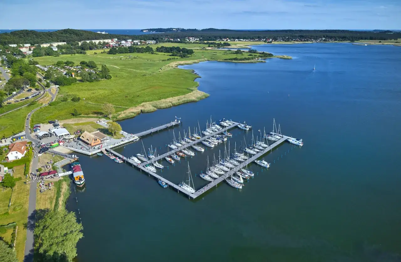 Der Hafen Sellin ist voller Segelboote, die im ruhigen Wasser liegen. Im Hintergrund erstreckt sich eine grüne Landschaft mit Häusern und Bäumen.