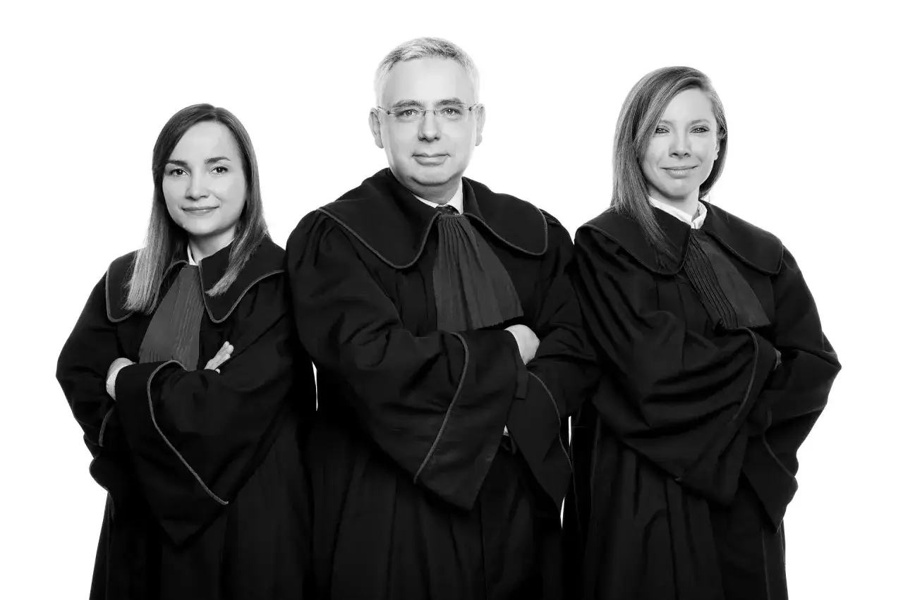 Trzy osoby w togach prawniczych, dwie kobiety i mężczyzna, stoją na białym tle.
