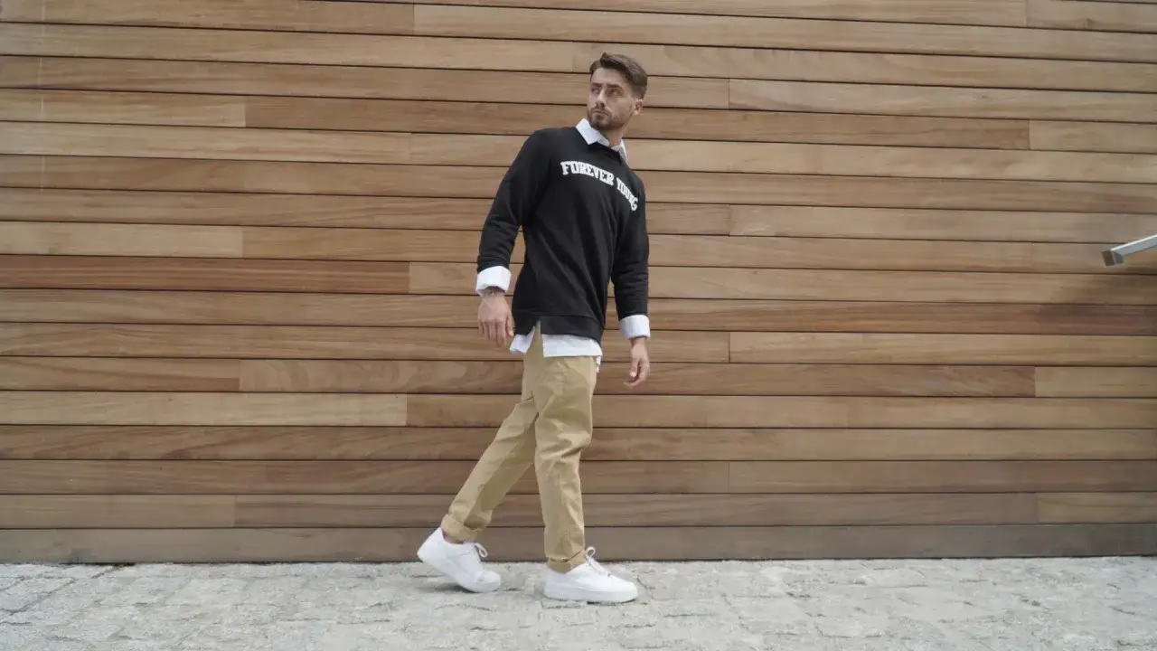 Mężczyzna w stylu smart casual, w beżowych spodniach i czarnej bluzie z białym napisem, na tle drewnianej ściany.