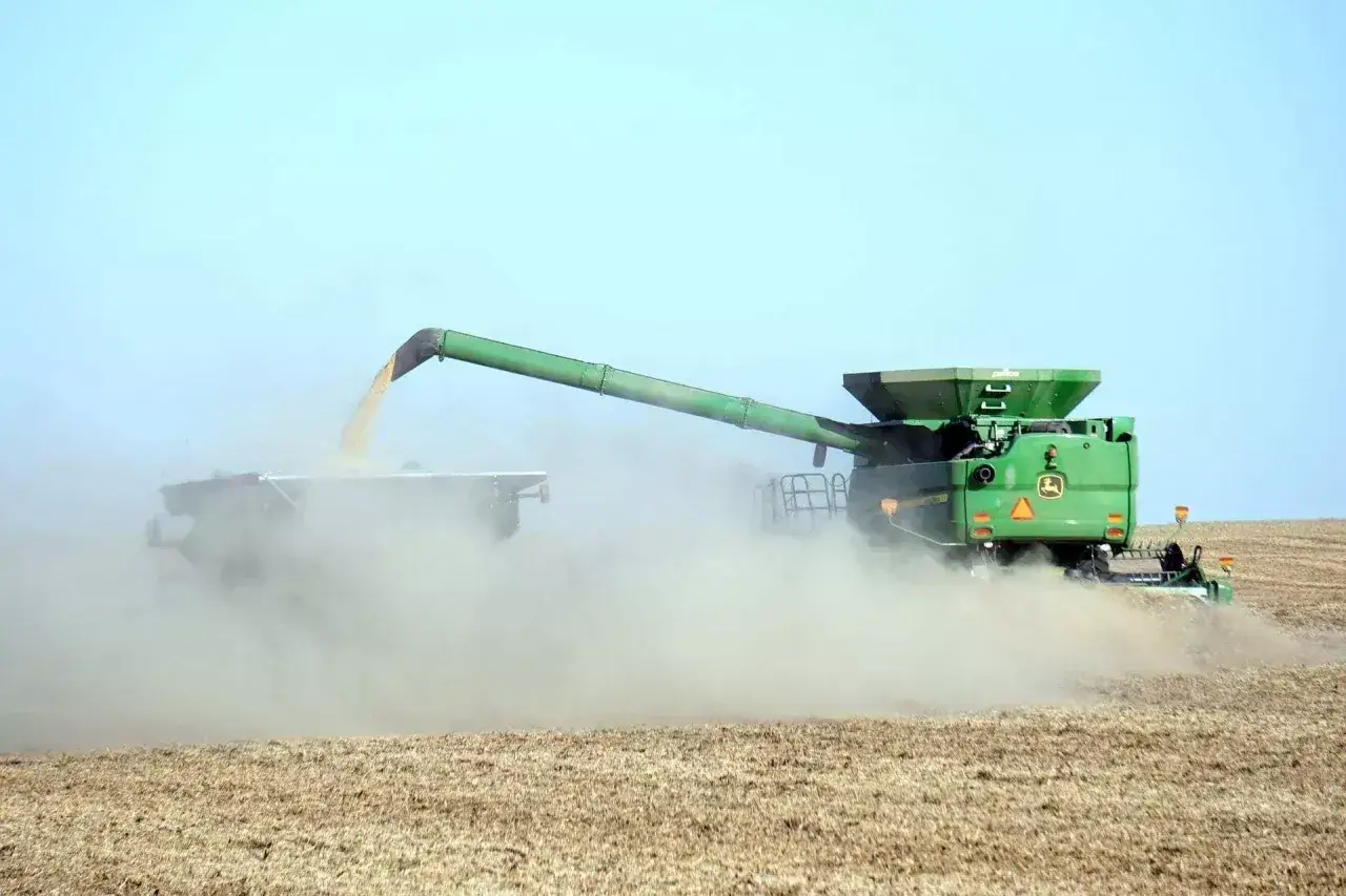 Kombajn John Deere podczas żniw, wysypujący pszenicę do przyczepy. Widać kurz unoszący się z pola, co sugeruje duży plon i pytanie: ile pszenicy z hektara?
