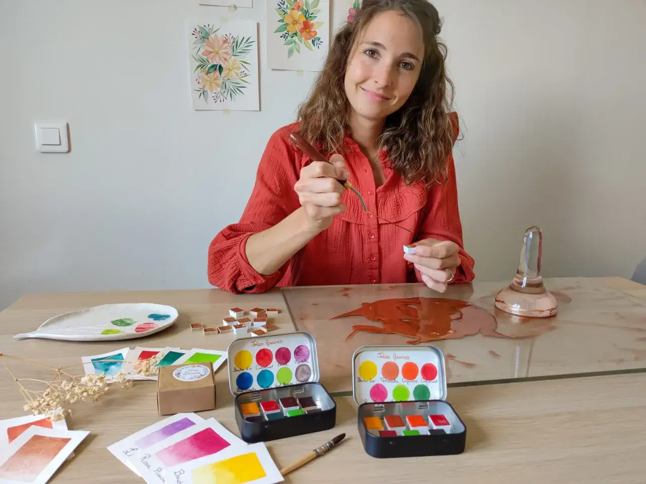Une artiste pr&eacute;pare des couleurs pour apprendre comment faire de l'aquarelle maison. Des palettes, des pastilles de couleur et des &eacute;chantillons sont &eacute;tal&eacute;s sur la table.
