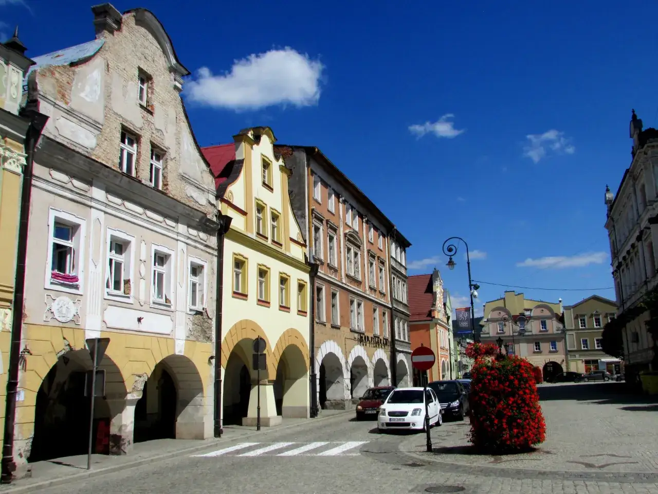 Kolorowy rynek w Lądku Zdroju z arkadowymi podcieniami kamienic i kwitnącą czerwienią.