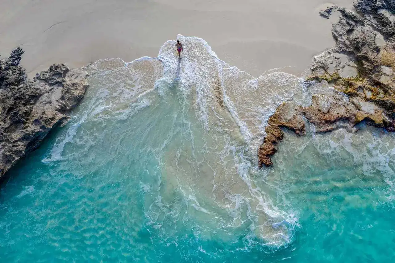 Frau genießt einen Badeurlaub im Winter am einsamen Strand, umspült von türkisfarbenem Wasser und Felsen.