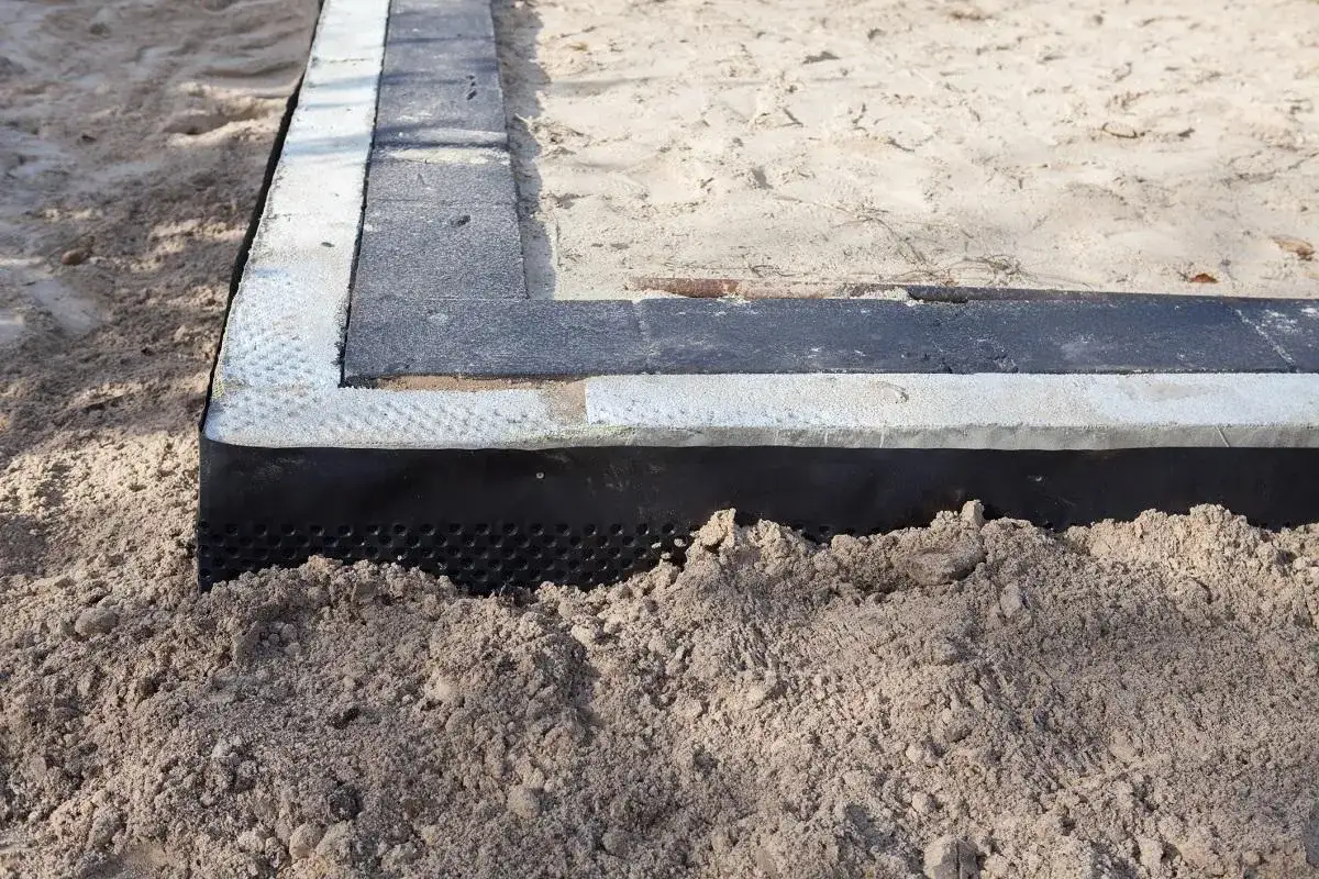 Warstwa folii izolacja pozioma chroni fundament przed wilgocią, otoczona piaskiem i elementami konstrukcyjnymi.