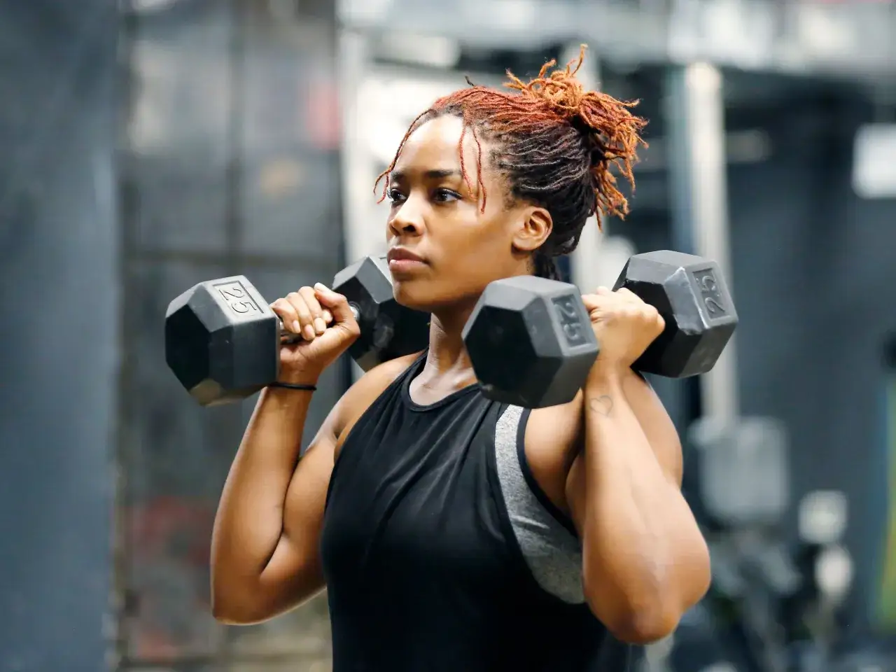 Frau mit roten Dreadlocks hebt Hanteln. Ihr Fokus liegt auf dem Muskelaufbau, sie trainiert konzentriert im Fitnessstudio.