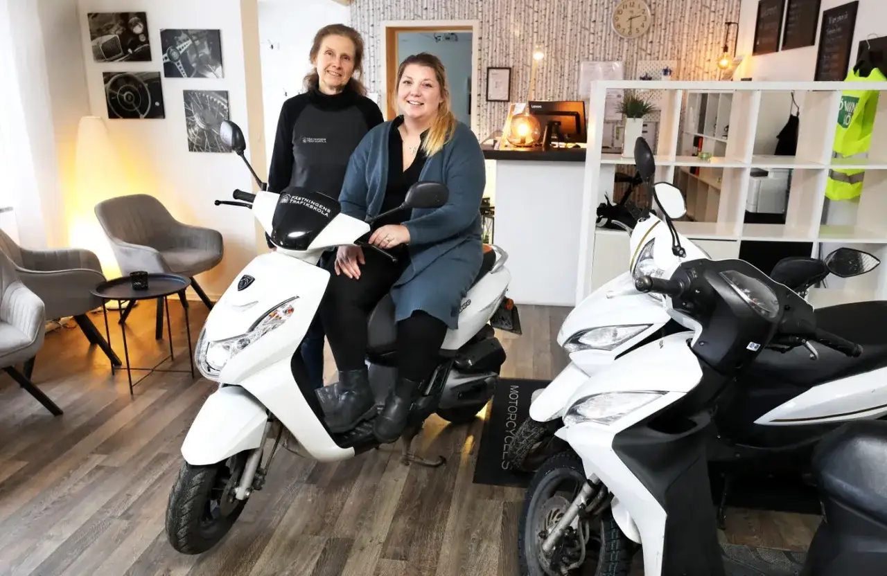Två kvinnor sitter på en vit moped. Kanske de diskuterar mopedkort pris och hur man får det.