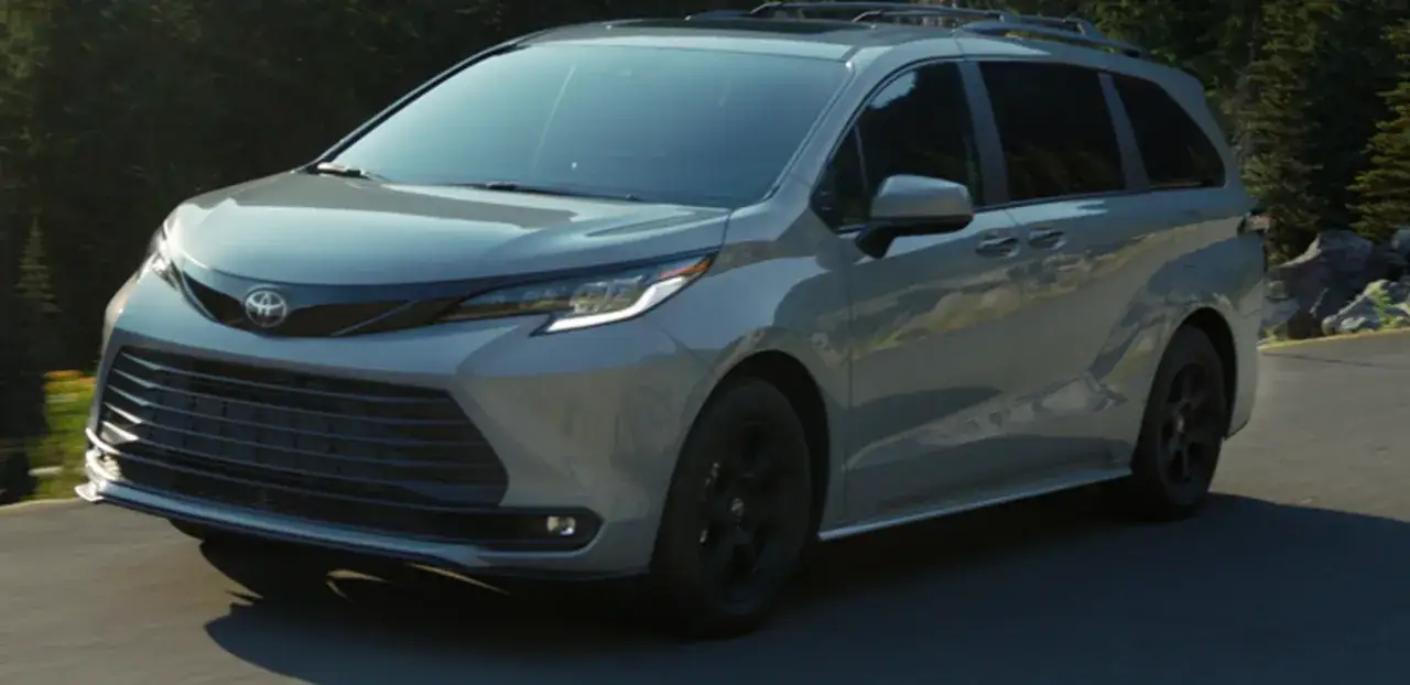 Srebrna Toyota Sienna z czarnymi felgami jedzie po krętej drodze wśród drzew.