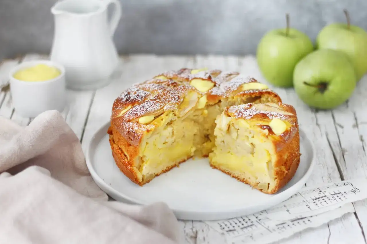 Torta di mele con crema pasticcera, soffice e dorata, cosparsa di zucchero a velo. Accanto, mele verdi fresche e una ciotolina di crema.