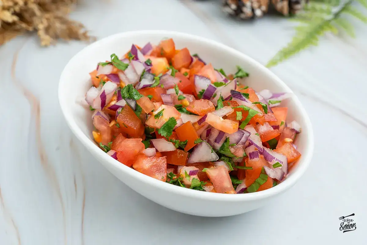 Un tazón blanco lleno de pico de gallo, con sus vibrantes ingredientes: tomate, cebolla morada y cilantro picados.
