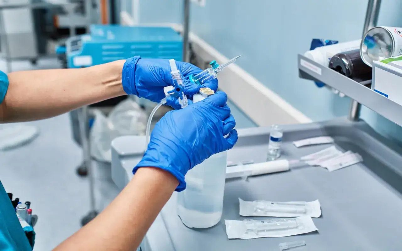 Hände mit blauen Handschuhen bereiten eine Infusion vor, indem sie eine Spritze an einen Infusionsbeutel anschließen.
