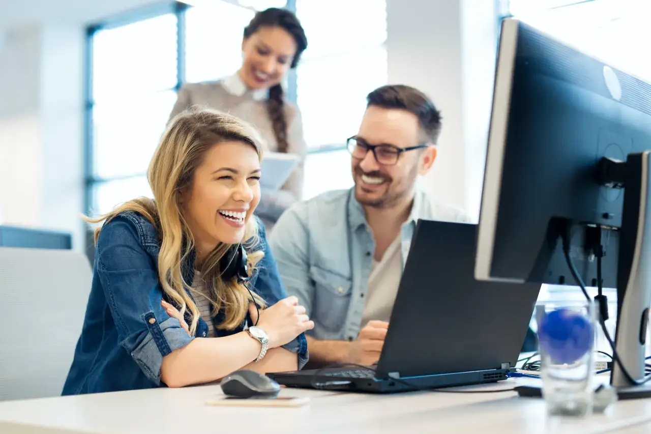 Uśmiechnięci pracownicy przy biurku, śmiejący się z czegoś na laptopie. W tle koleżanka też się uśmiecha. Wszyscy mają dobre postojowe w pracy.