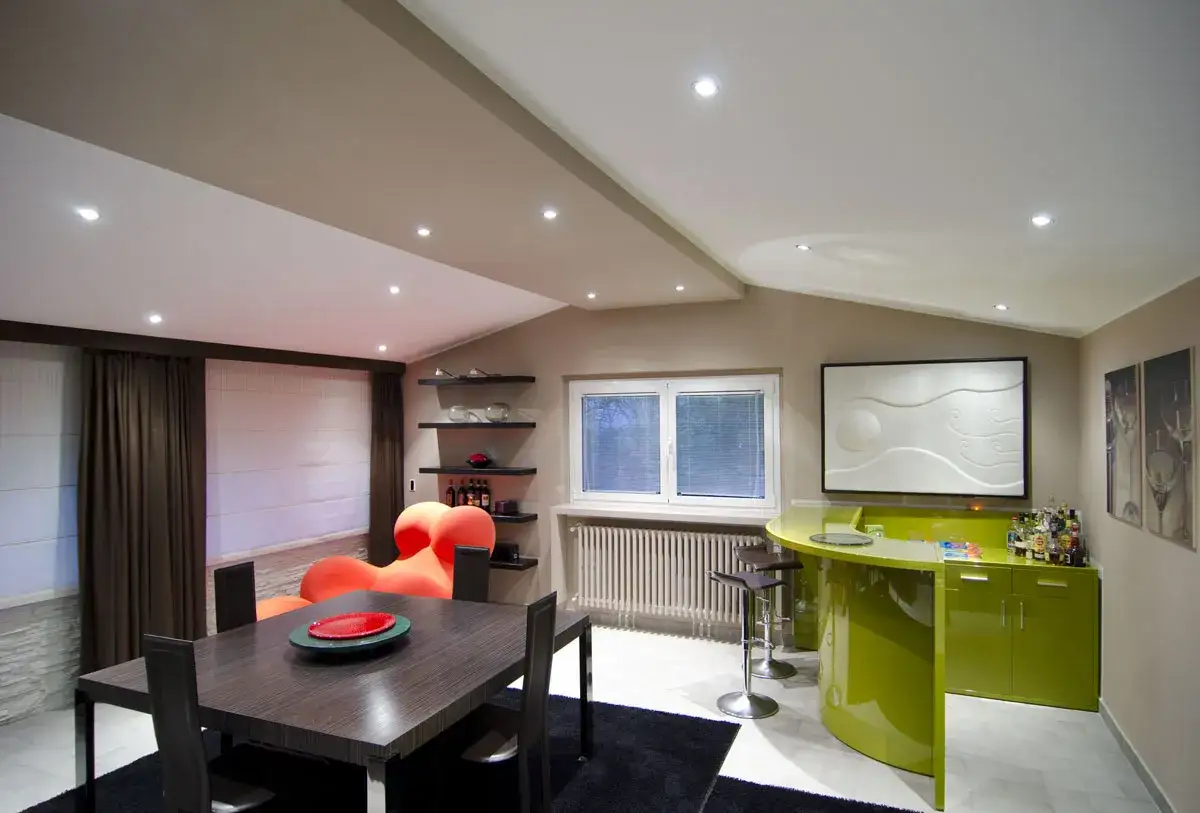 Soggiorno moderno con tavolo da pranzo, divano arancione e bancone bar verde brillante. Il soffitto a più livelli, con un interessante spessore, è illuminato da faretti.