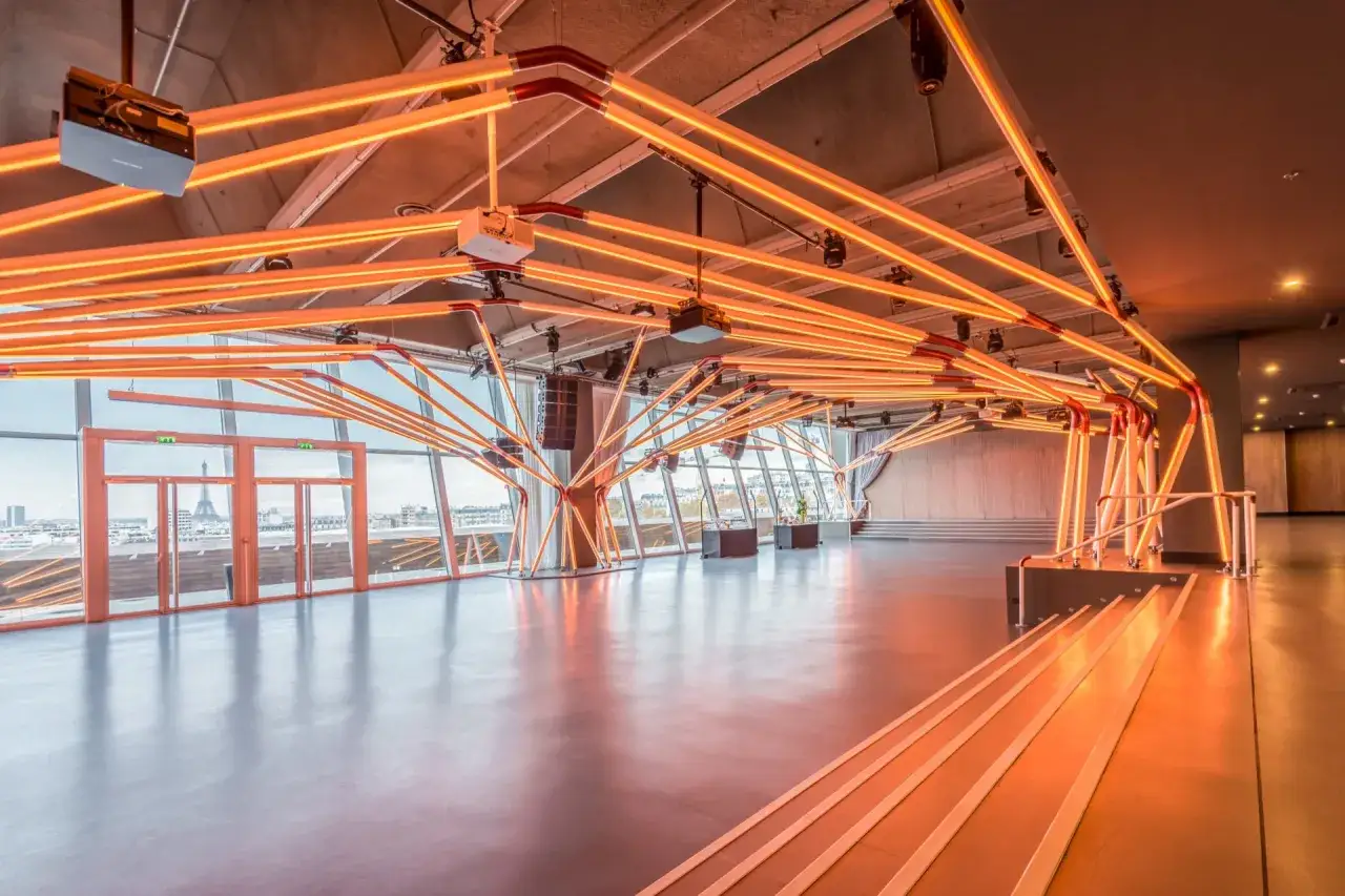 Un espace événementiel moderne à Paris, illuminé par des néons orange vifs. La Tour Eiffel est visible à travers les grandes baies vitrées.