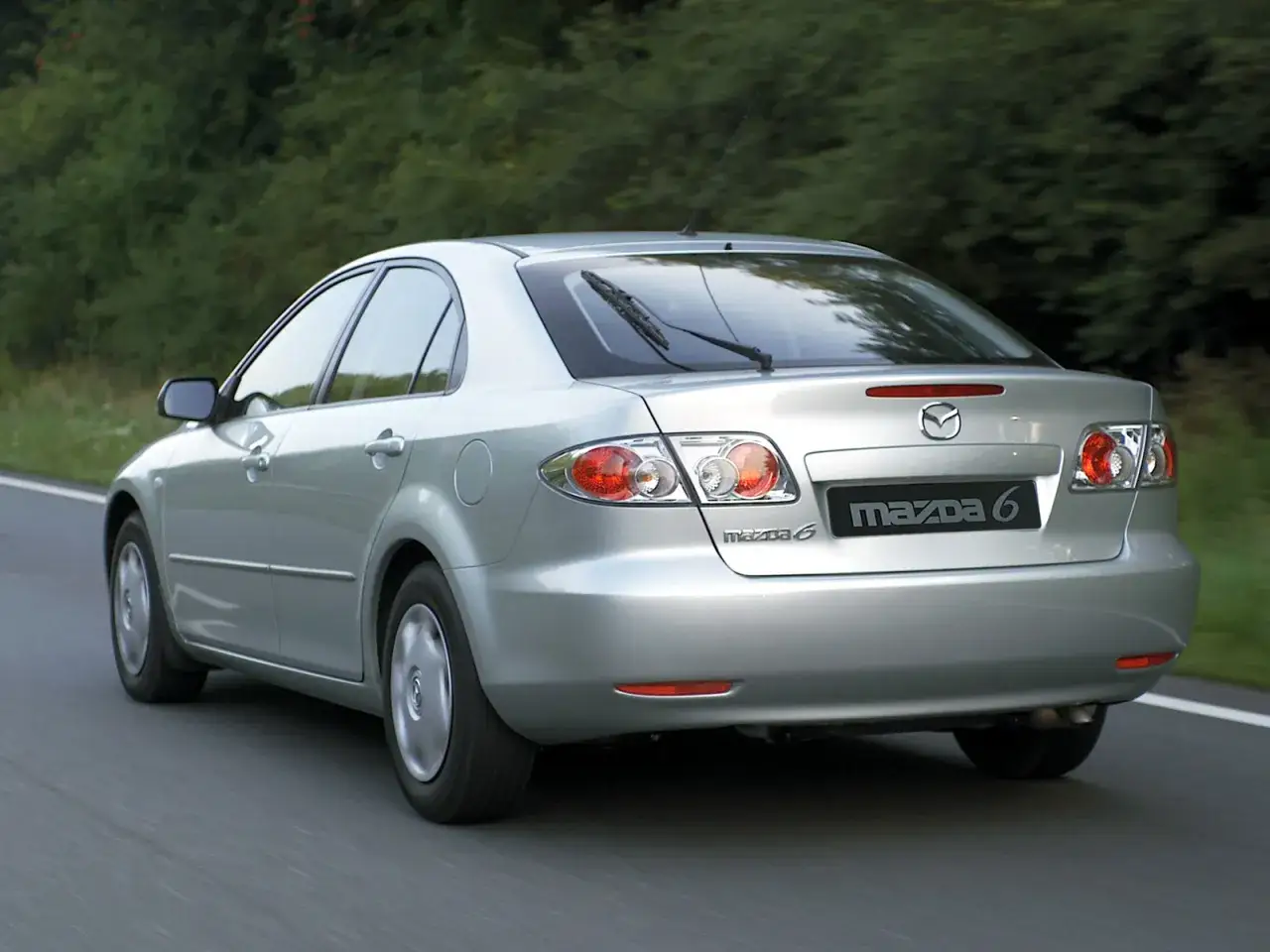 Srebrna Mazda 6 GG jedzie po drodze, otoczona zielenią.