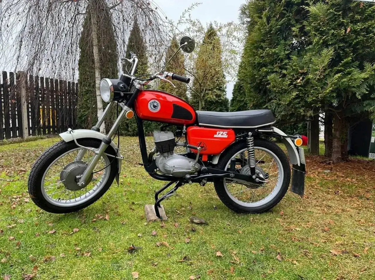 Klasyczny, czerwony motocykl WSK 125 stoi na trawie, otoczony drzewami.