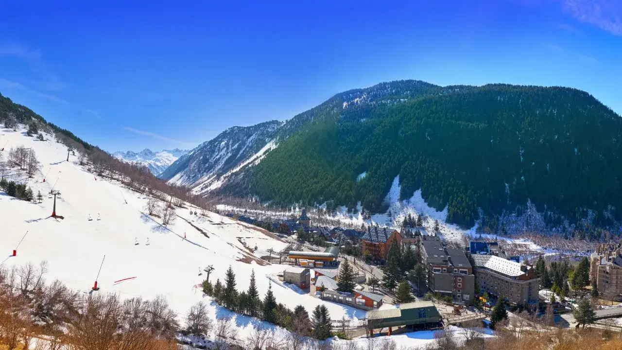 Paisaje invernal en el **Valle de Arán en autocaravana**. Montañas nevadas, pistas de esquí y un pueblo alpino bajo un cielo azul.