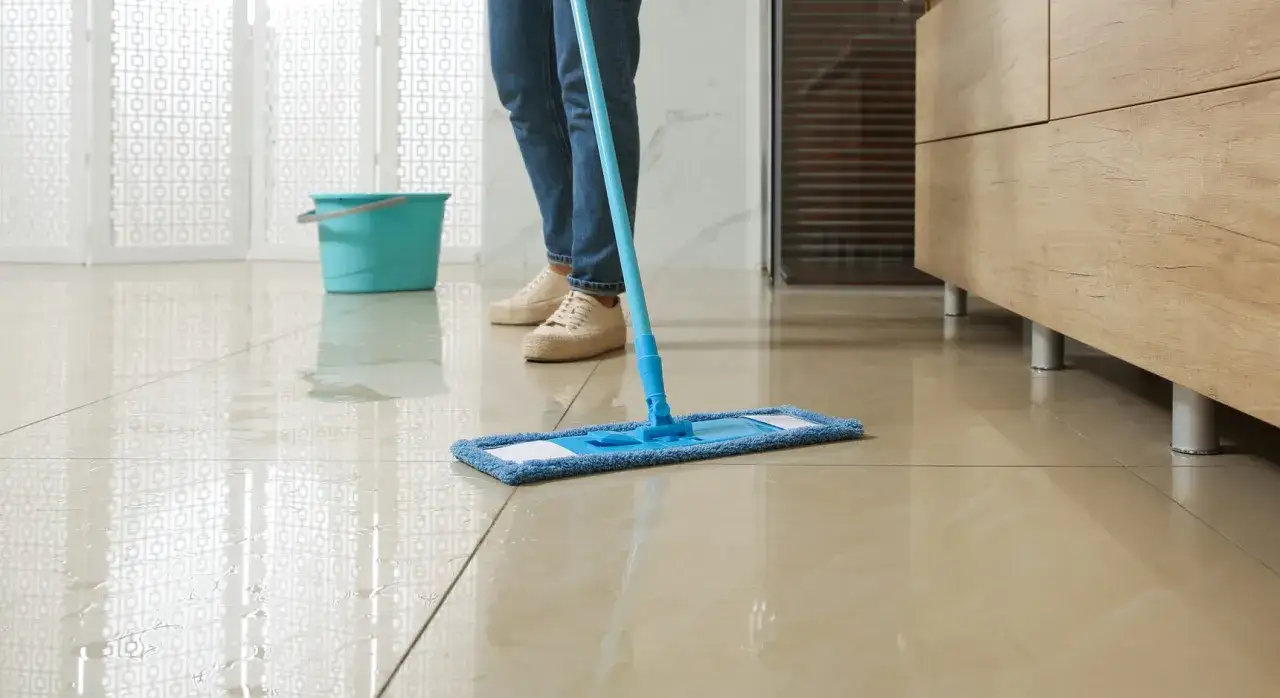 Person wischt mit einem blauen Wischmopp über einen nassen Marmorboden. Ein Eimer steht daneben.