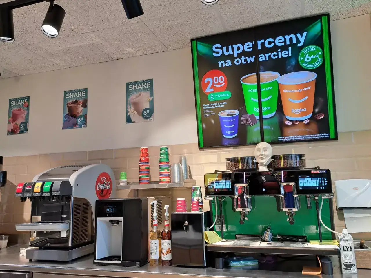 Kawa i napoje w Żabce, w tym bezkofeinowa, na ekranie z promocją. Obok dystrybutor Coca-Coli i ekspres do kawy.