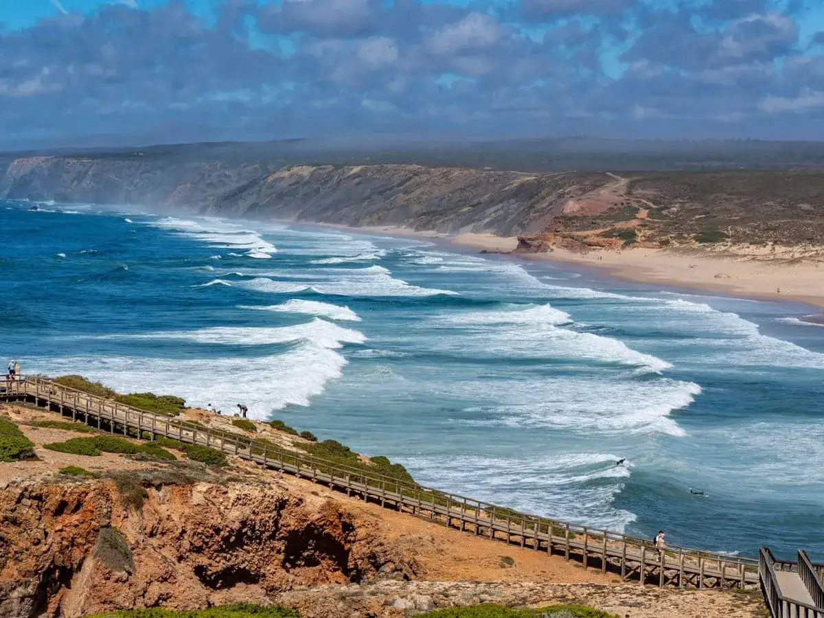 Holzsteg am Fischerweg Portugal führt zu einem weiten Sandstrand mit tosenden Wellen. Surfer genießen die Brandung.