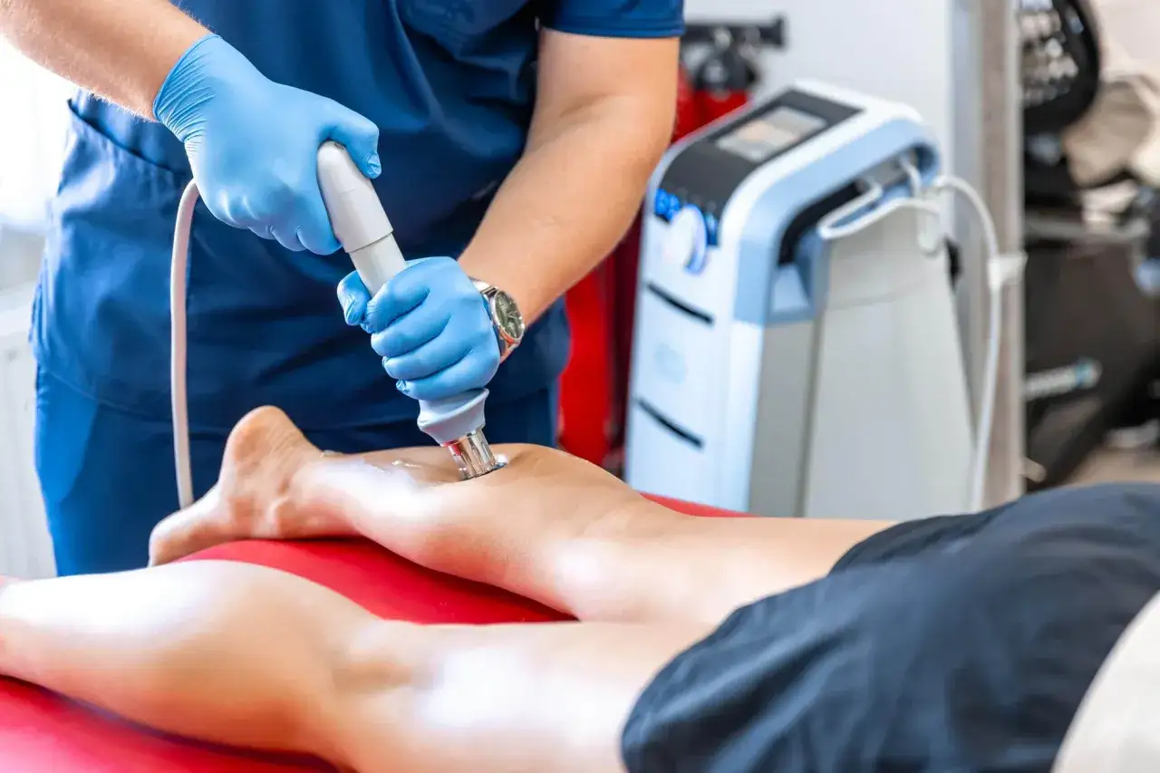 Fizjoterapeuta w niebieskich rękawiczkach wykonuje zabieg falą uderzeniową na łydce pacjenta. Nowoczesne urządzenie wspiera rehabilitację.