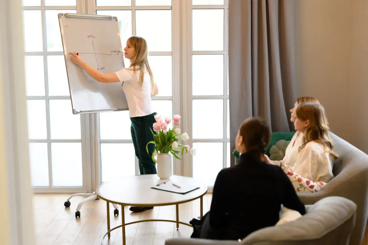 Psychiatra z autyzmem prowadzi sesję terapeutyczną, rysując na tablicy. W sali siedzą pacjenci.
