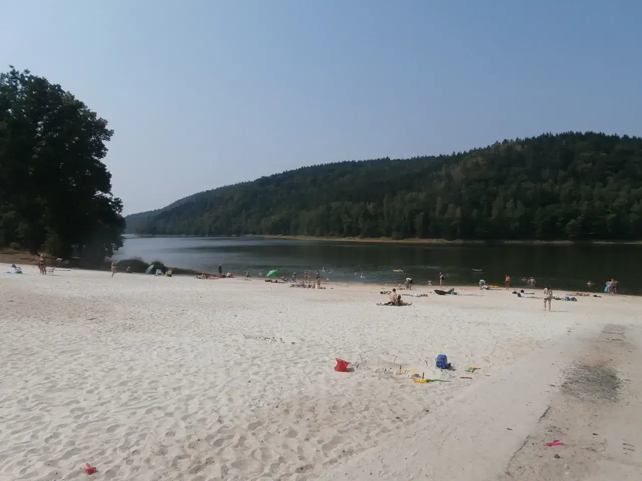 Plaża nad jeziorem, ludzie odpoczywają na piasku i kąpią się w wodzie. To idealne miejsce na wypoczynek, jak na **złoty potok camping**.