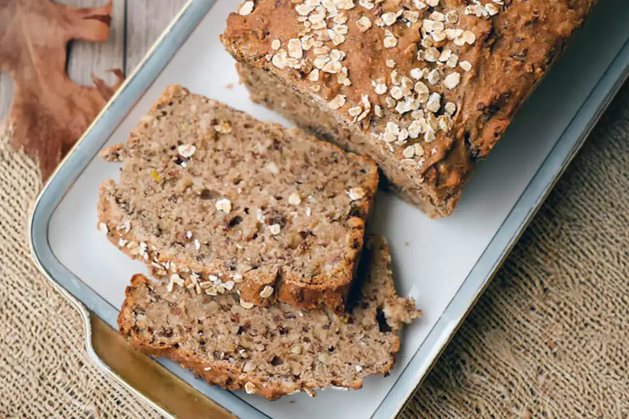 Ein rustikales Vollkornbrot in Kastenform, mit Haferflocken bestreut, liegt auf einem Teller. Zwei Scheiben sind bereits abgeschnitten.