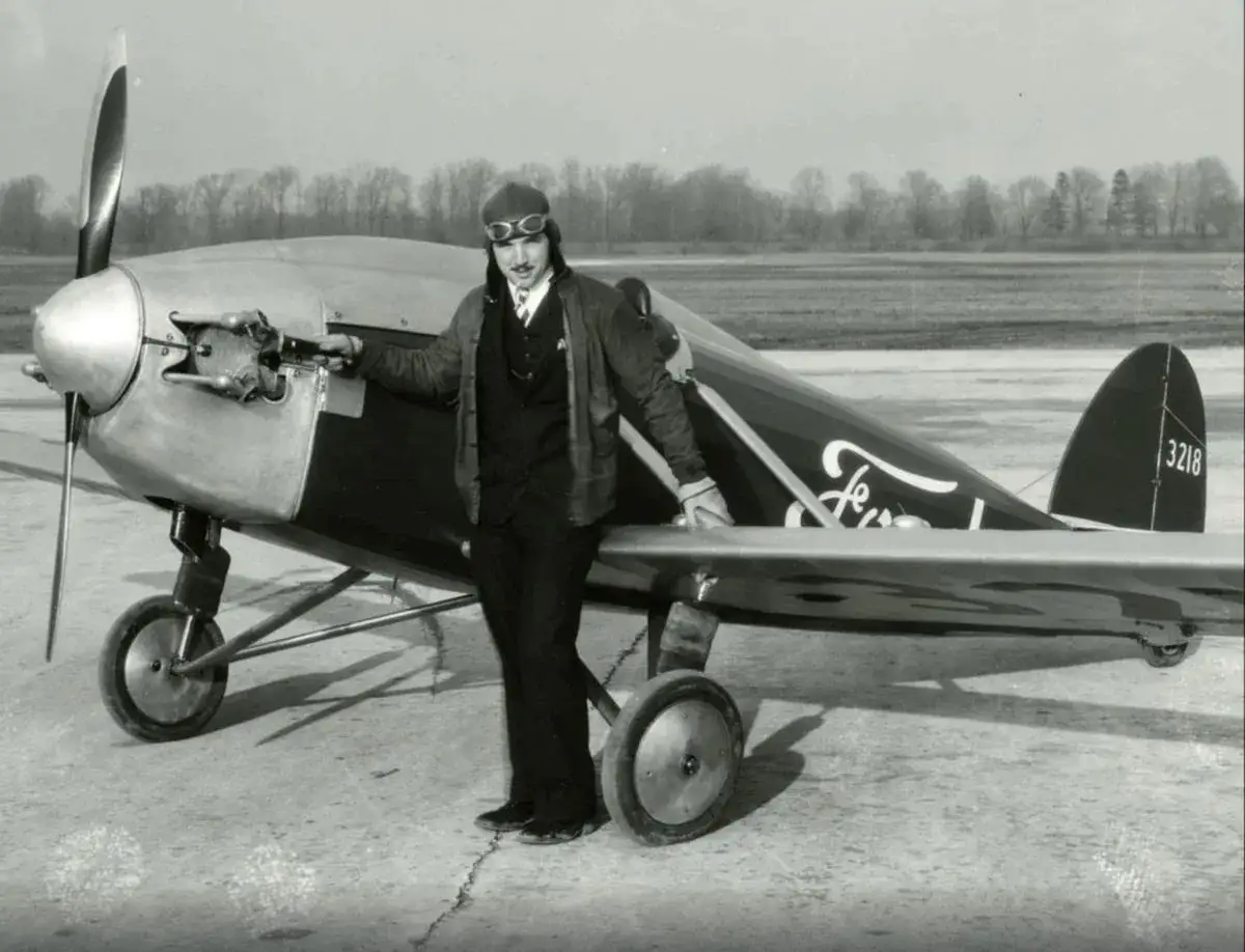 Ein Pilot in Fliegerkleidung steht neben einem Ford Flugzeug. Das Flugzeug hat einen Propeller und Räder.