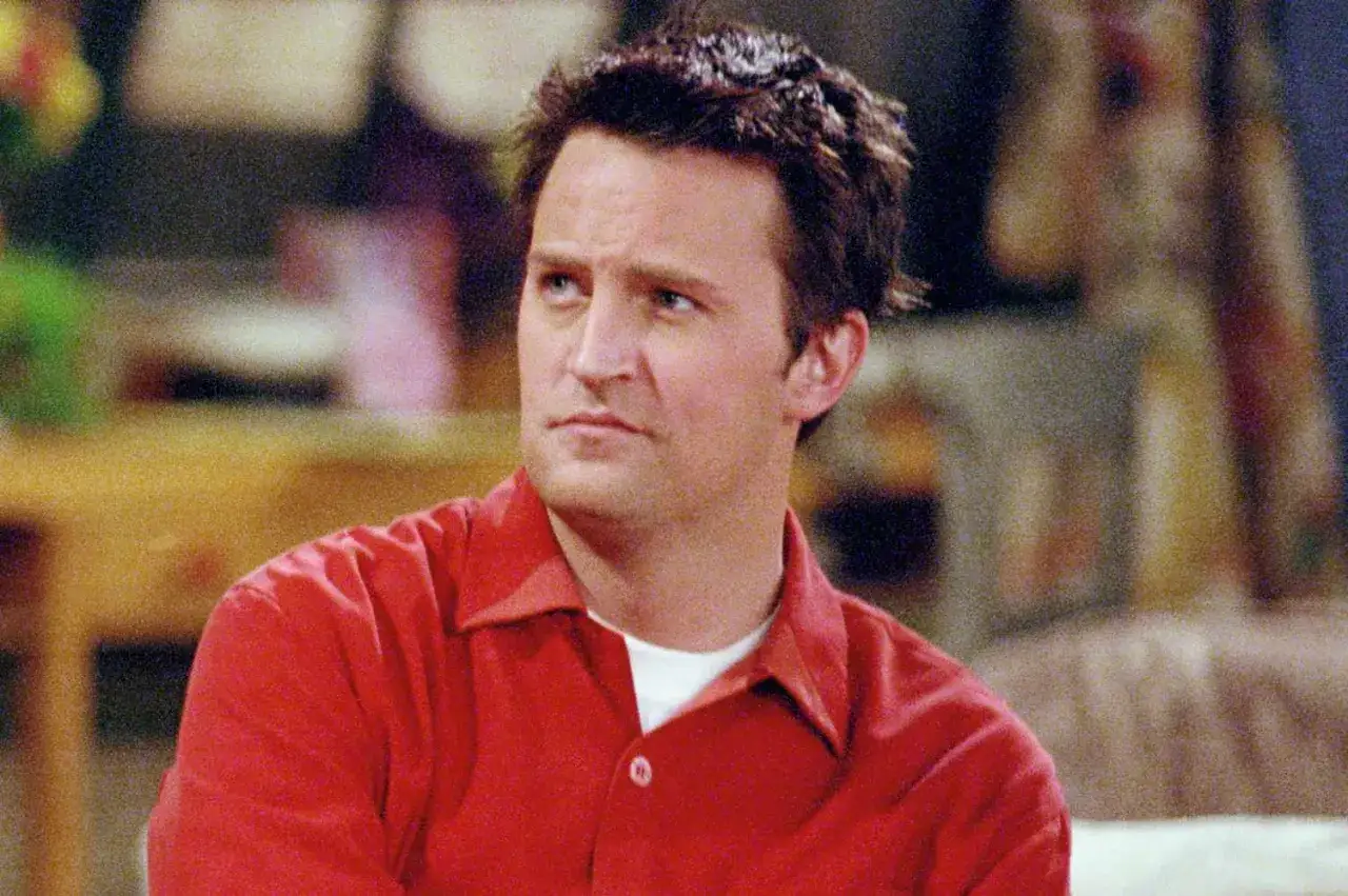 Chandler Bing w czerwonej koszuli, zamyślony, w tle widać fragmenty mieszkania.