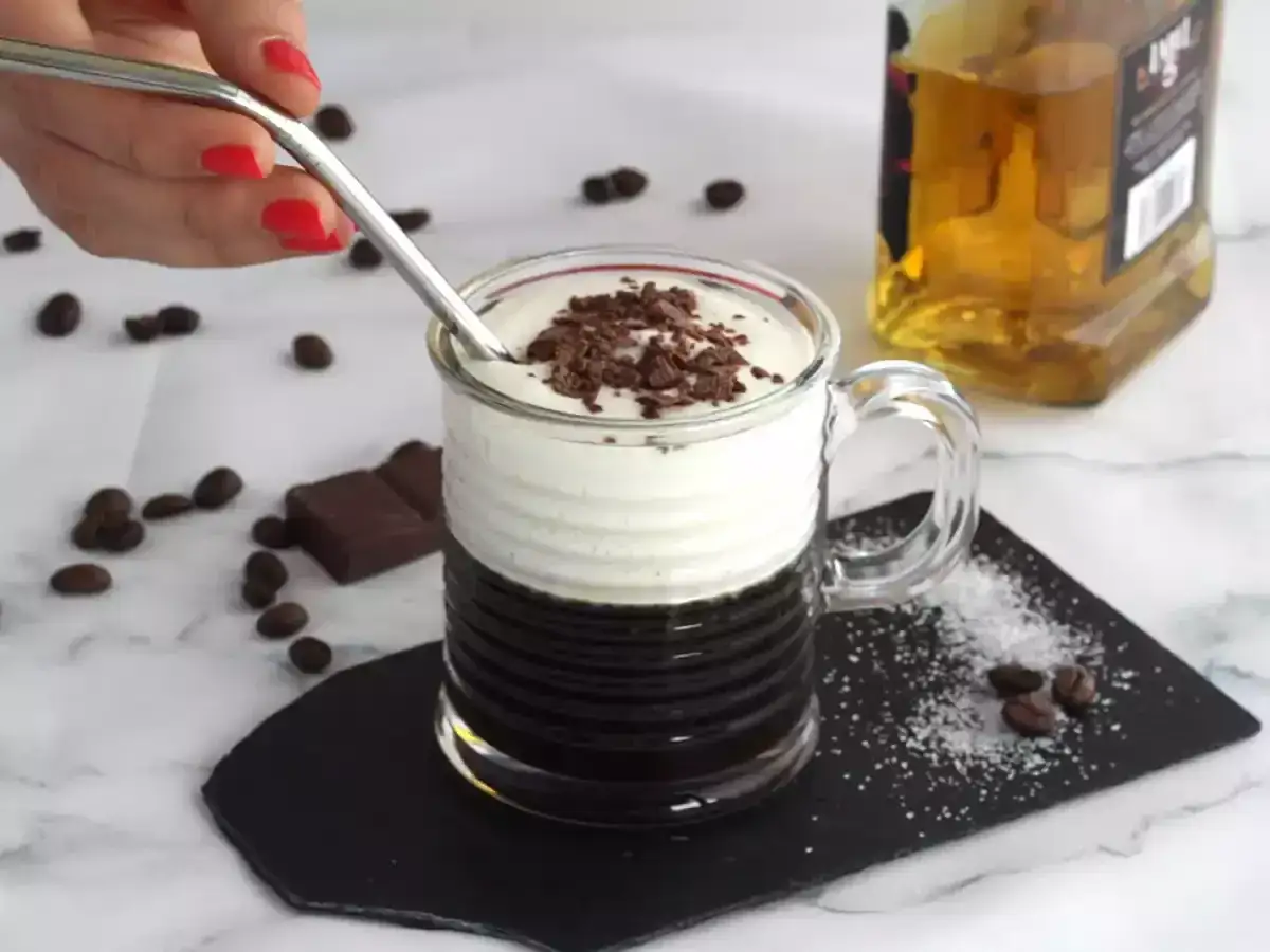 En hand med r&ouml;d nagellack r&ouml;r i en Irish coffee med vispgr&auml;dde och chokladstr&ouml;ssel. Bredvid st&aring;r en flaska whiskey.