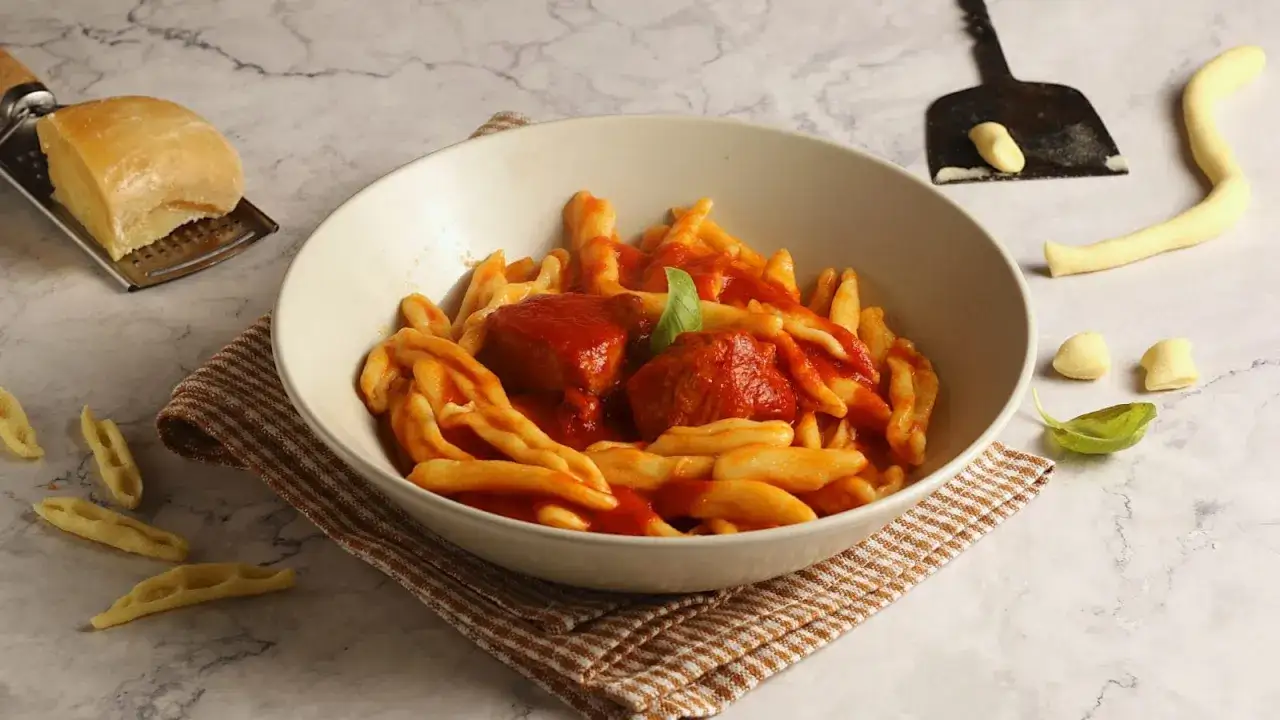 Un piatto di strascinati pugliesi con sugo rosso e polpette, accanto a formaggio grattugiato e pasta cruda.