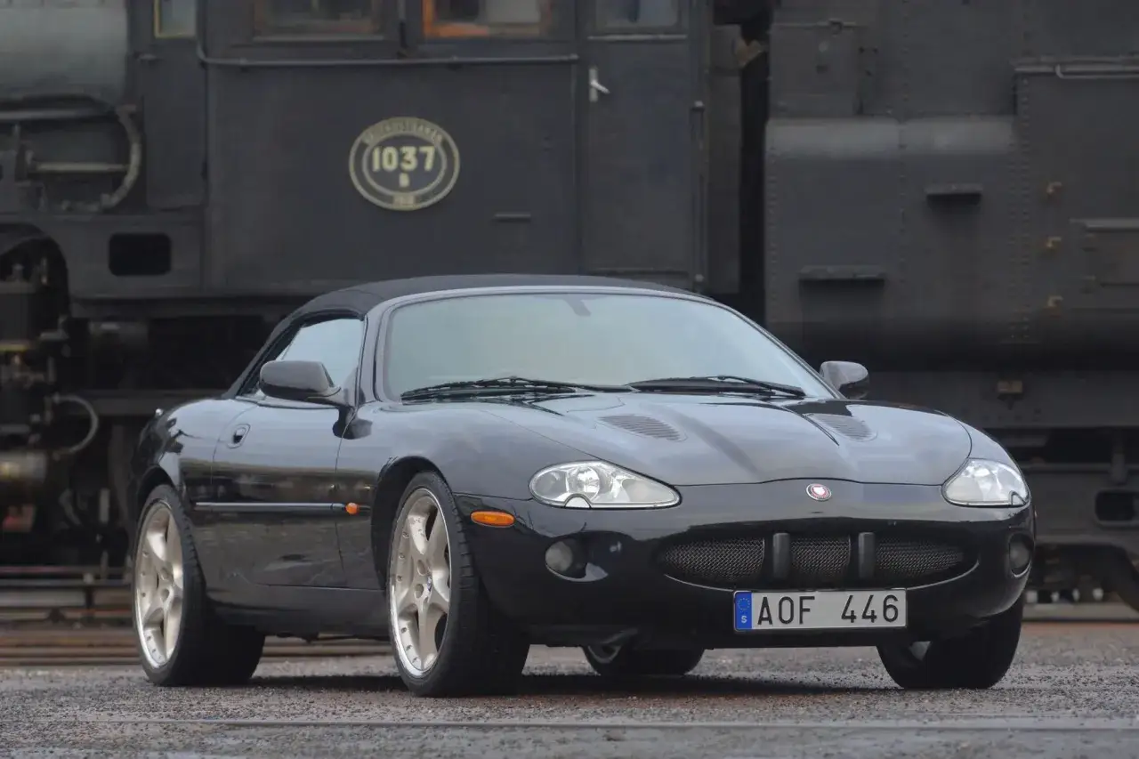Svart Jaguar XKR till salu, en klassisk sportbil med taket nere, parkerad framf&ouml;r ett gammalt &aring;nglok.