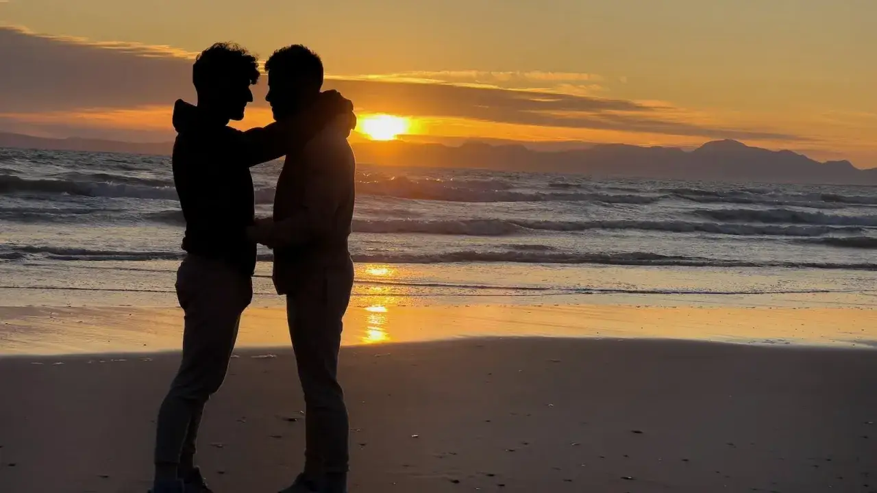 Silhouetted couple embracing on a beach at sunset, a perfect moment on their gay honeymoon.