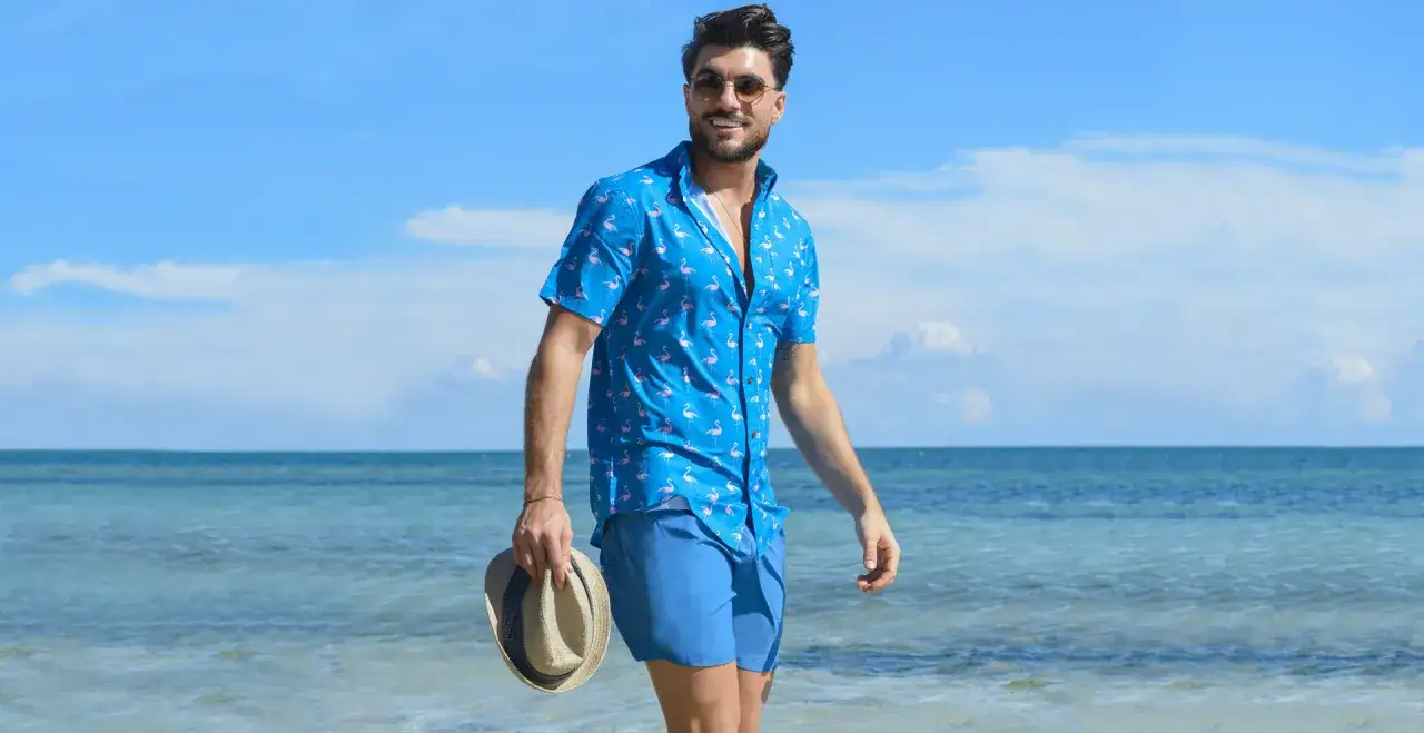 Mężczyzna w niebieskiej koszuli w flamingi i szortach, w kapeluszu, na plaży. Styl hawajski.