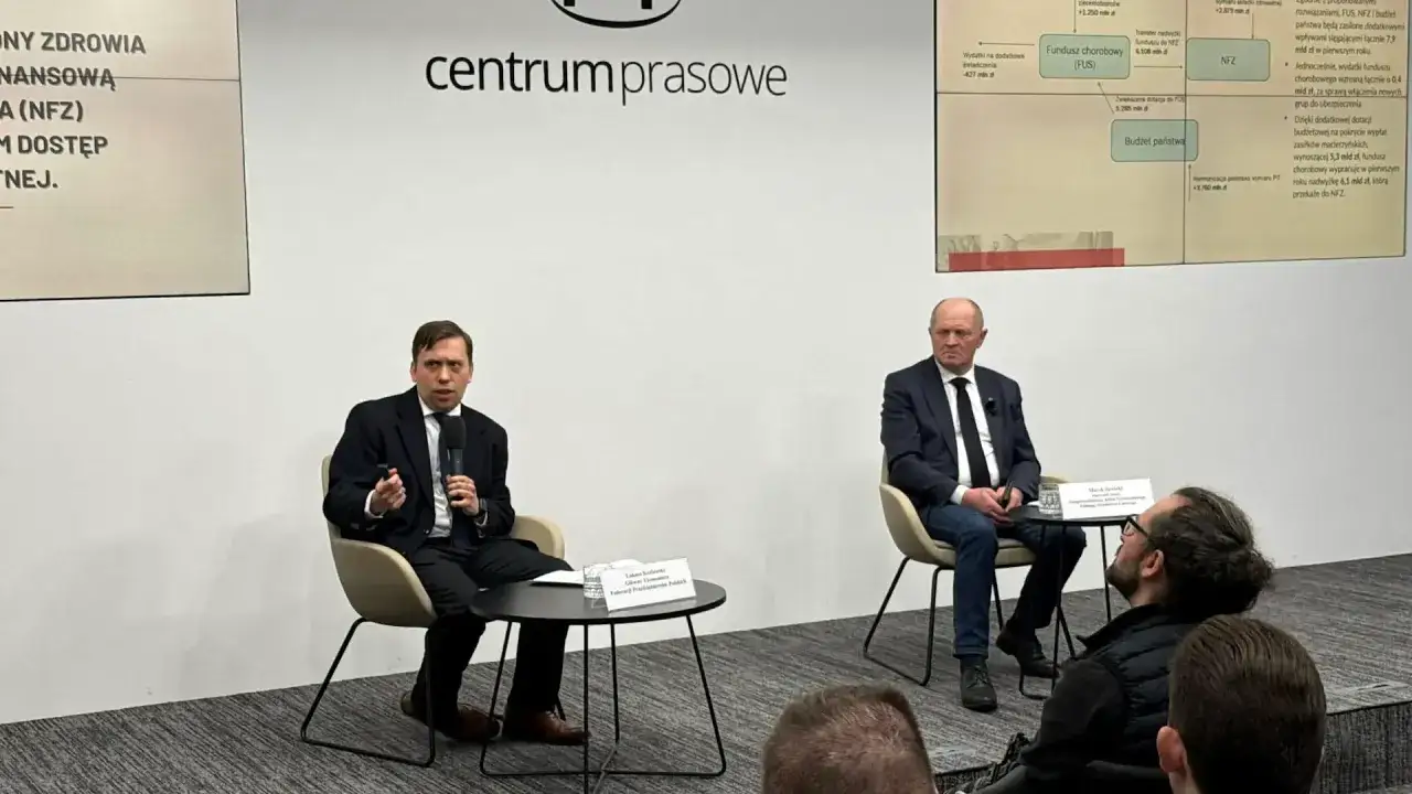 Dyskusja o tym, dlaczego PiS chce zwiększenia nakładów na ochronę zdrowia o 23 mld zł. Dwóch mężczyzn w garniturach siedzi na krzesłach, jeden mówi do mikrofonu.