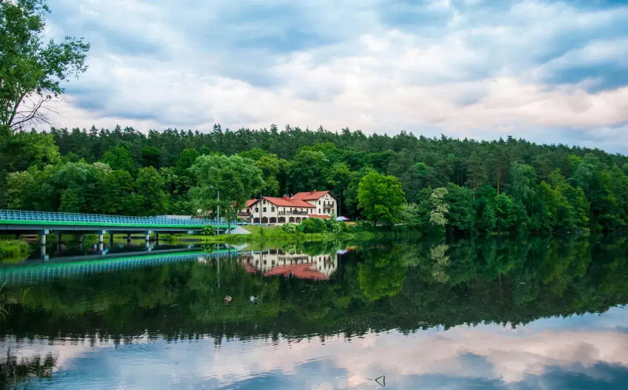 Malowniczy hotel nad jeziorem, z mostem i lasem w tle. Idealne miejsce na rodzinne wakacje, oferujące atrakcje dla dzieci.