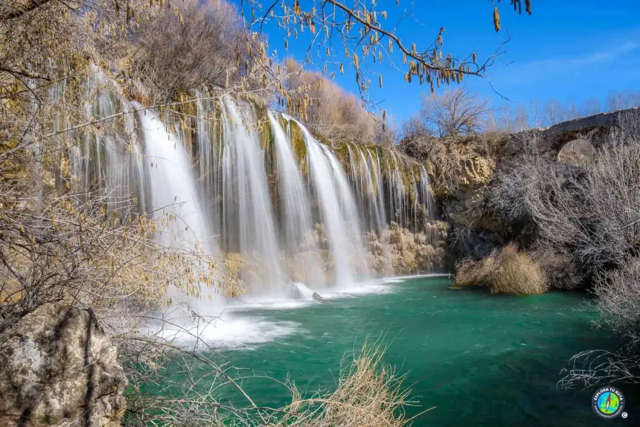 Cascada de agua turquesa, como los ojos del Cabriel, cayendo sobre rocas y rodeada de vegetaci&oacute;n seca bajo un cielo azul.