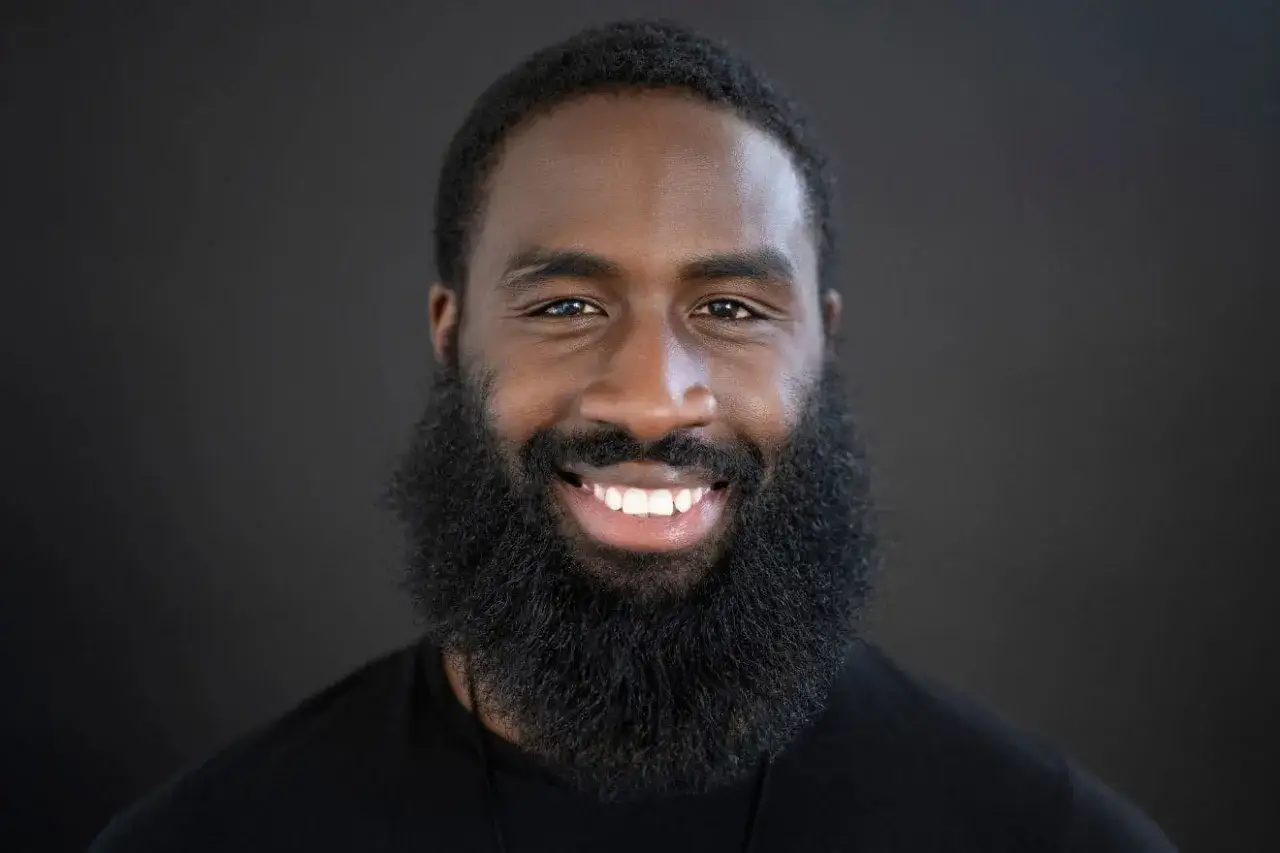 Hombre sonriente con una barba espesa y oscura, luciendo un atuendo negro.