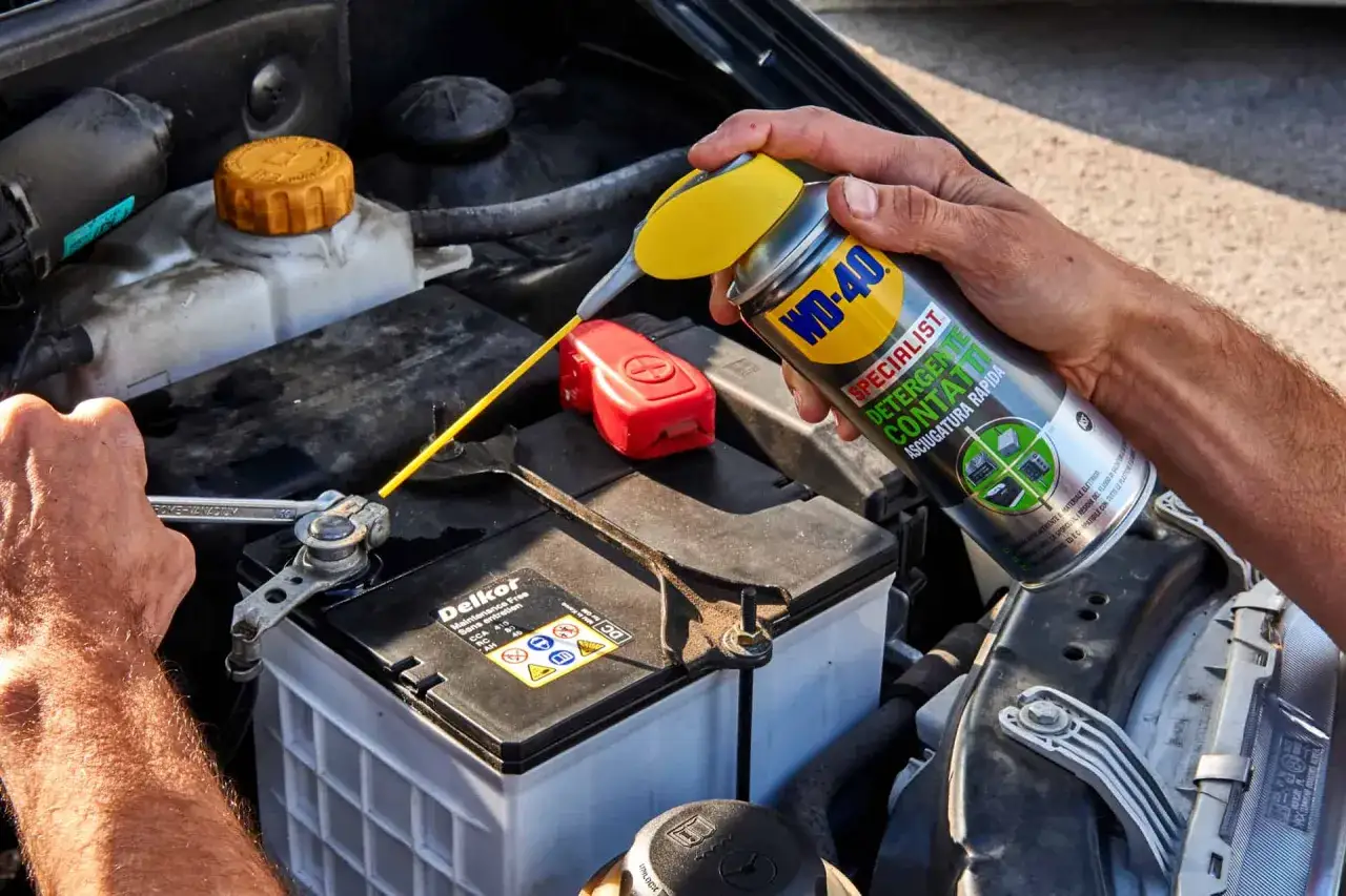 A hand sprays WD-40 Specialist onto car battery terminals, cleaning them for better connection.