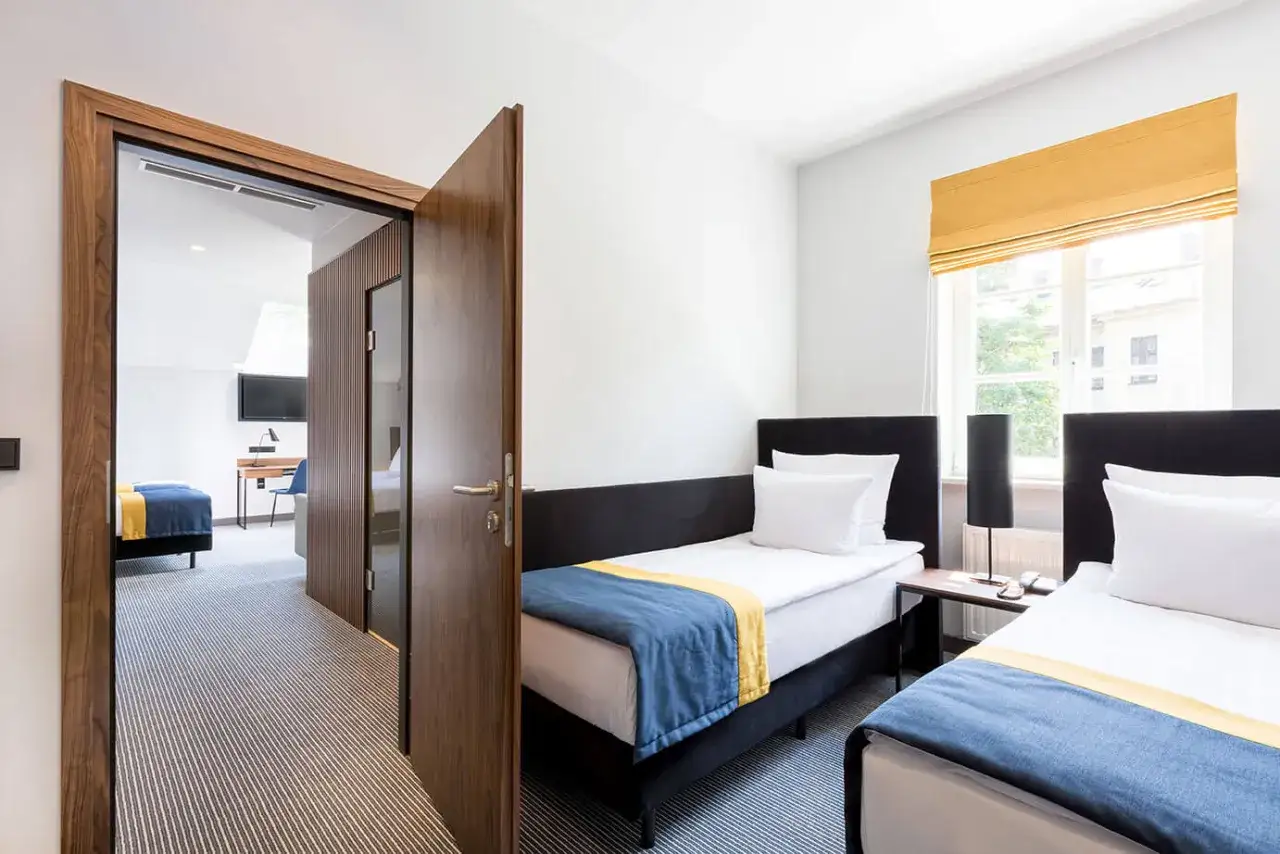 Dwa łóżka w apartamencie typu suite, z niebieskimi i żółtymi narzutami. W tle widać drugie pomieszczenie z łóżkiem i biurkiem.