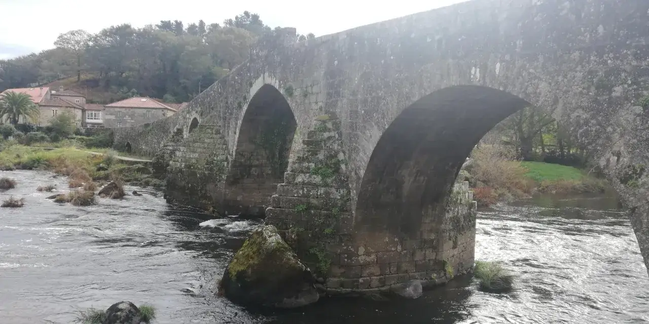 El puente Maceira, con sus arcos de piedra y el río que fluye, se alza majestuoso entre la vegetación y las casas del pueblo.