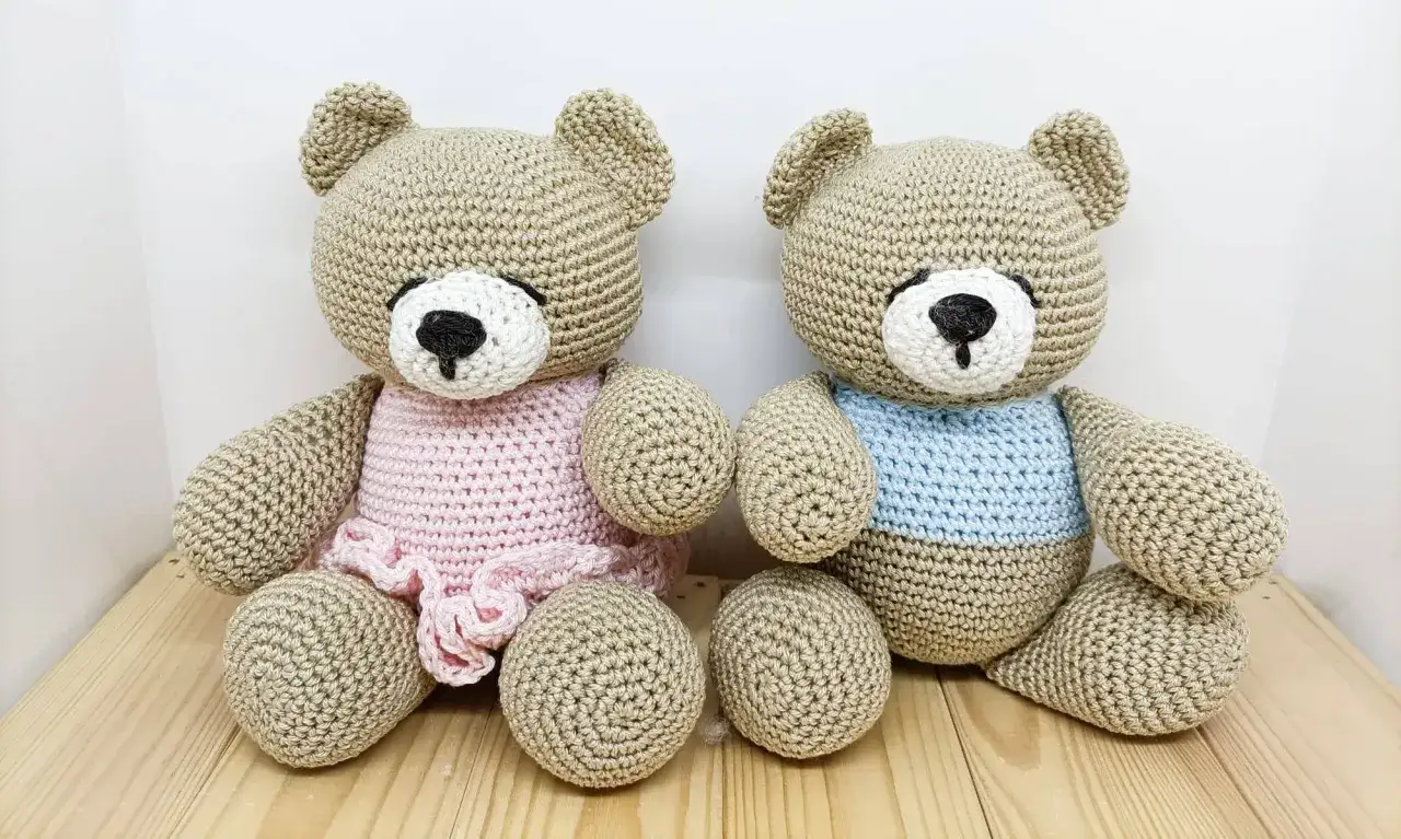 Due orsetti teddy amigurumi, uno con un vestitino rosa e l'altro con una maglietta azzurra, siedono fianco a fianco su un tavolino di legno.