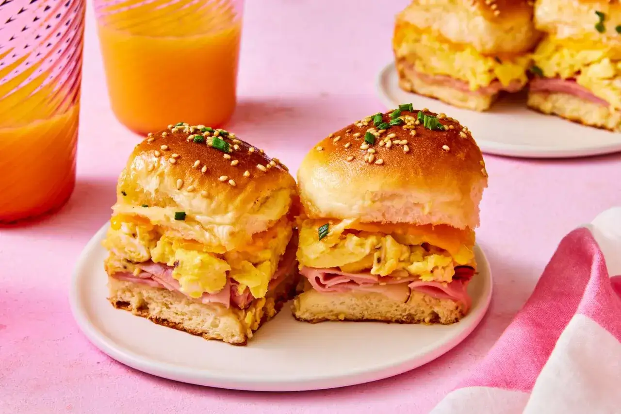 Zwei Frühstücks-Sliders Burger mit Rührei, Schinken und Käse auf einem Teller, daneben Orangensaft.