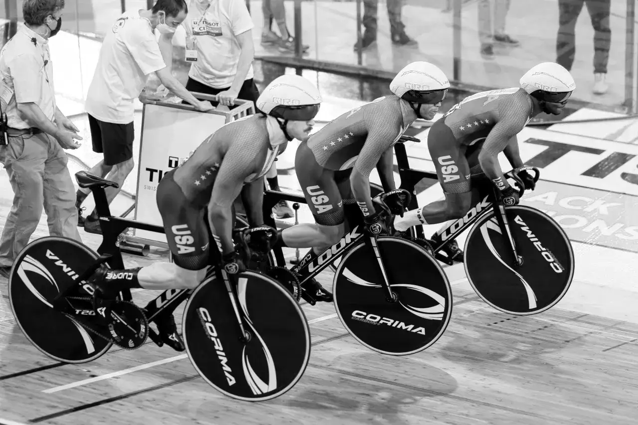 Three cyclists in USA kits prepare for a track sprint, helmets on, bikes poised.