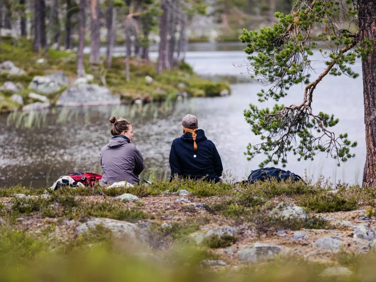 Tv&aring; personer sitter vid en sj&ouml; i skogen, redo f&ouml;r en eld brasa. Ryggs&auml;ckar ligger bredvid dem.