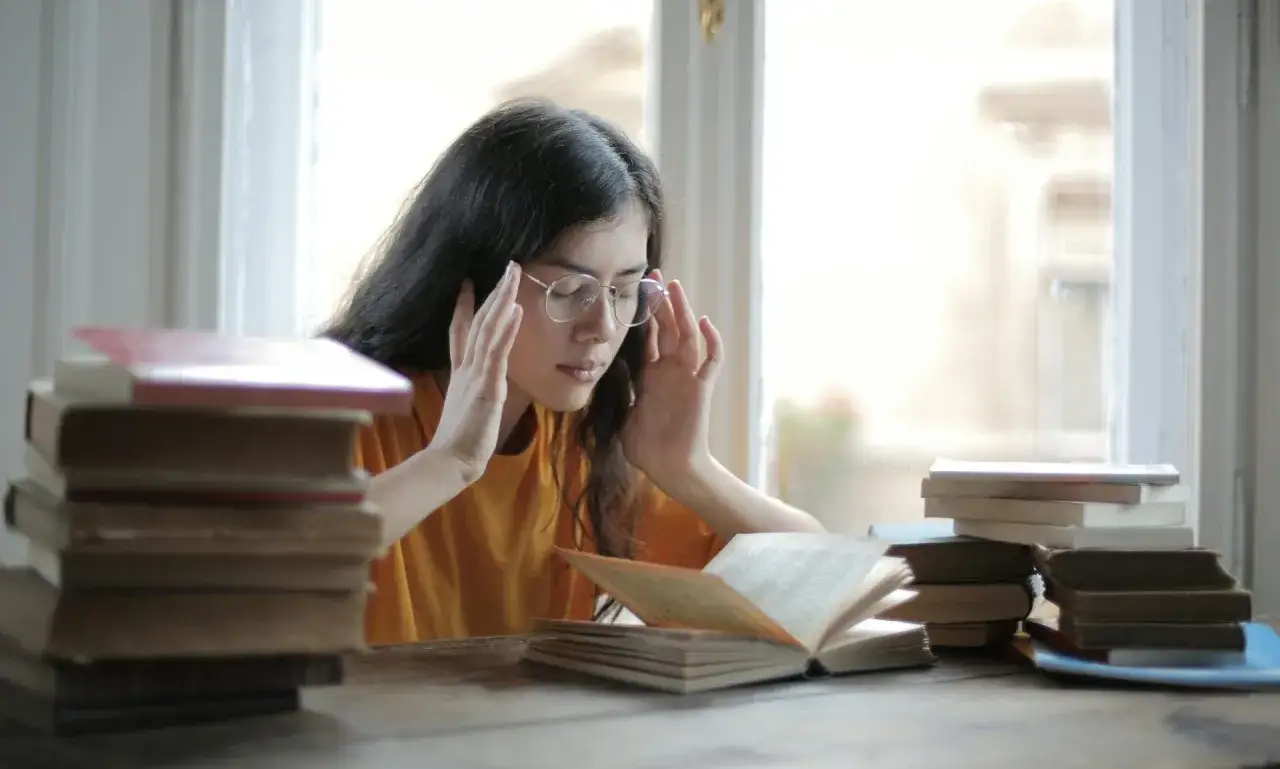 Zmęczona studentka z okularami, otoczona książkami, zastanawia się, ile wynoszą alimenty na studenta.