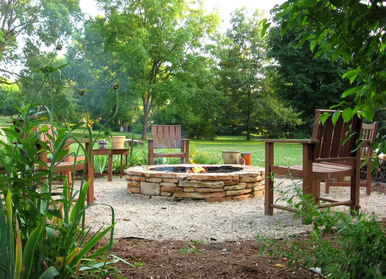 A stone fire pit with a fire burning, surrounded by gravel, wooden chairs, and lush greenery. This scene inspires thoughts on how to build a fire pit for cozy evenings.
