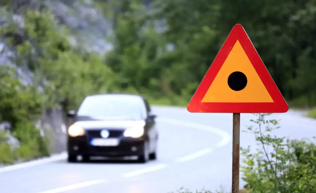 Se&ntilde;al de advertencia triangular con un punto negro en el centro, que indica que es un punto negro en la carretera. Un coche se acerca en la curva.