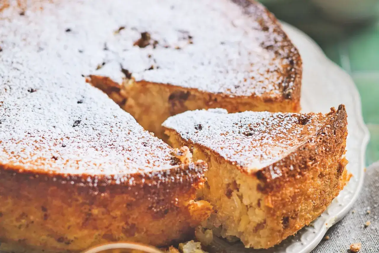 Torta di riso ricetta originale, soffice e dorata, cosparsa di zucchero a velo. Una fetta è già stata servita, invitando a gustare questo dolce tradizionale.